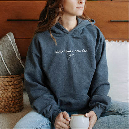 Person wearing a blue hoodie with text saying 'make Heaven Crowded' , sitting on a couch holding a mug.