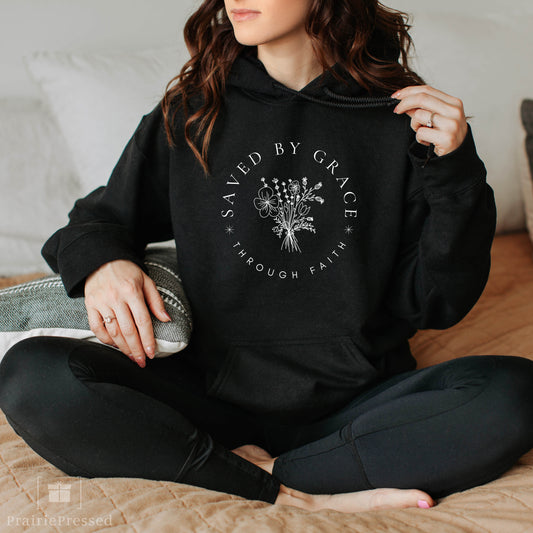 Person wearing a black hoodie with 'Saved by Grace Through Faith' text and floral design.
