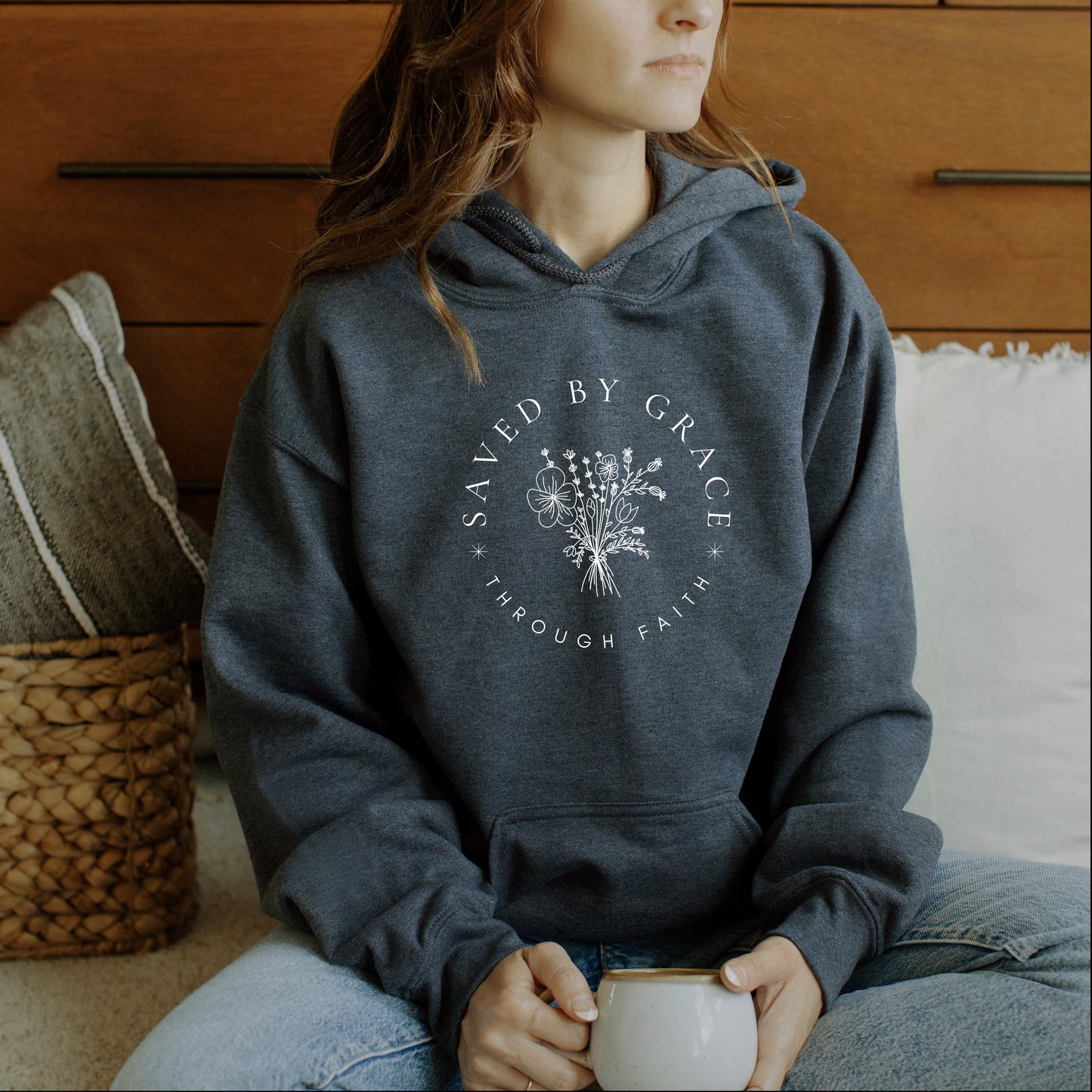 Young woman wearing a hoodie with 'Saved by Grace Through Faith' text and floral design, holding a mug.