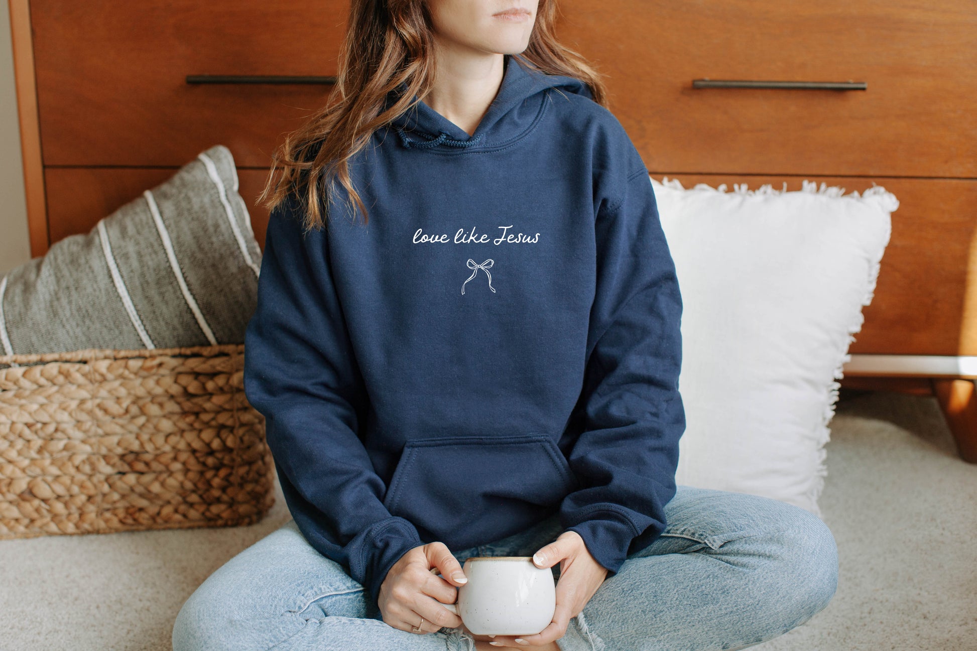 Person wearing a navy hoodie with text  "Love like Jesus" sitting on a couch holding a mug.